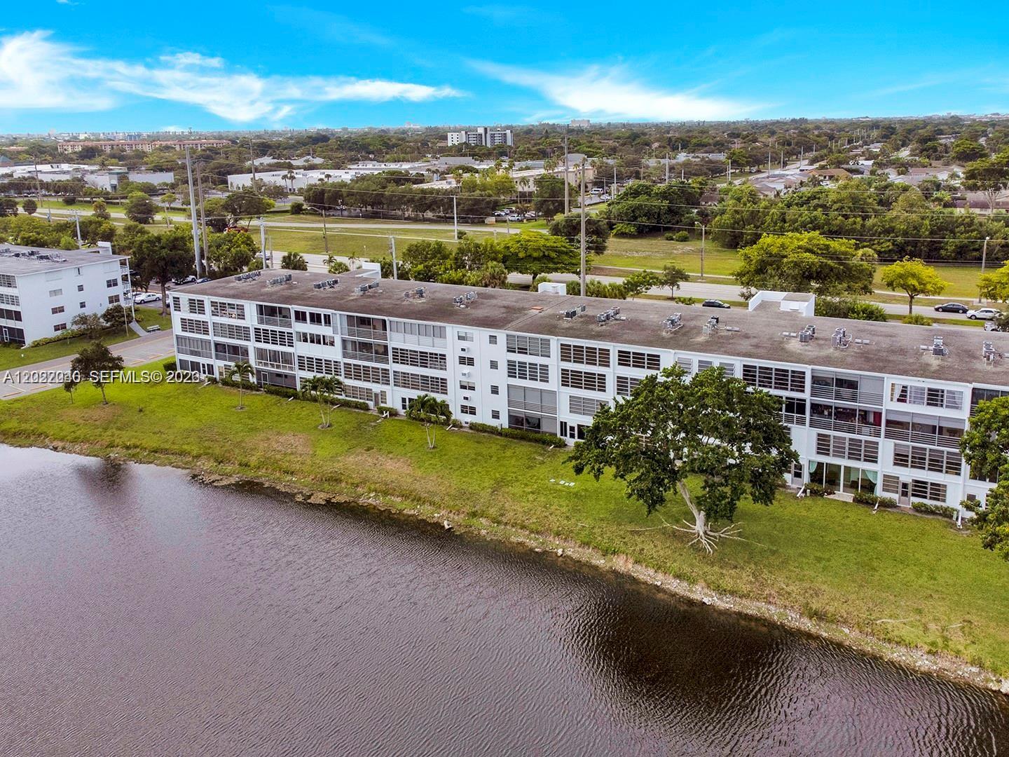 An aerial view shows a long, multi-story white apartment building situated beside a lake — 4052 E Harwood E # 4052, Deerfield Beach FL 33442, DEERFIELD BEACH, FL