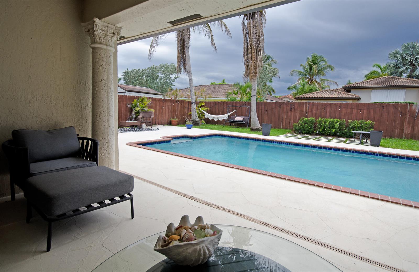 A covered patio overlooks a swimming pool in a backyard with palm trees and fencing — 18324 SW 149th Place, Miami FL 33187, MIAMI, FL