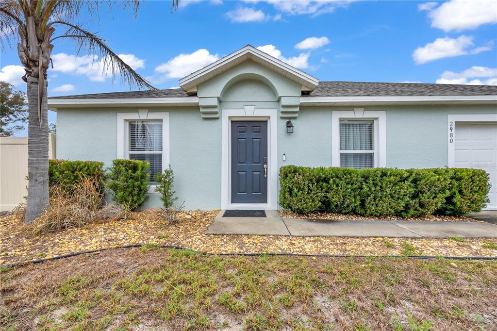 The exterior of a single-story house features a light green facade, a dark door, and landscaping — 2980 BEAL ST STREET, DELTONA, FL