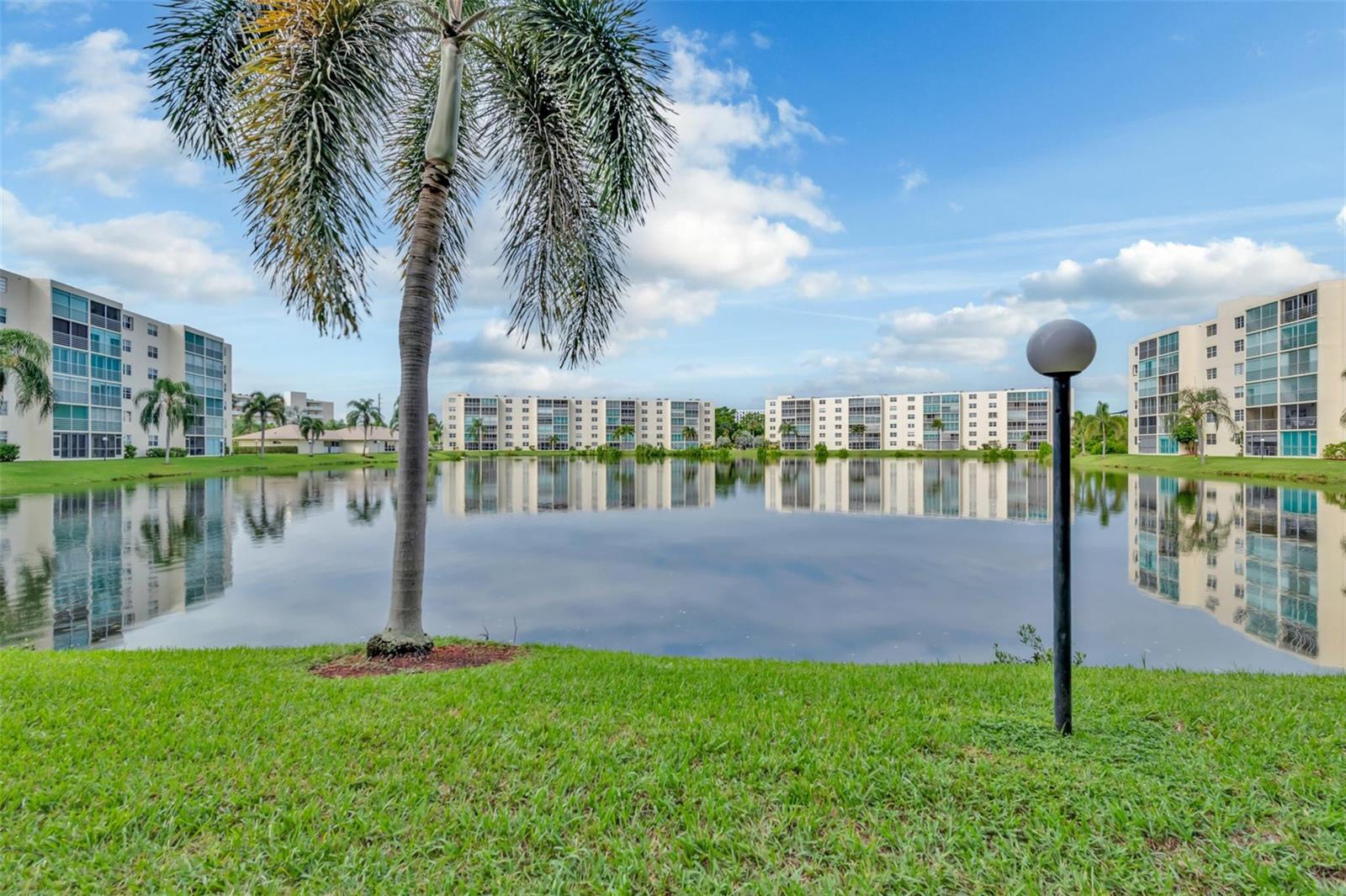 A calm lake reflects several multi-story apartment buildings surrounded by green lawns and palm trees — 121 SE 3rd Avenue # 104, Dania Beach FL 33004, DANIA BEACH, FL