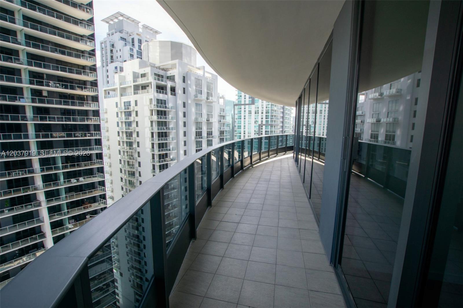 A curved balcony with tiled flooring and glass railings overlooks surrounding high-rise apartment buildings — 1000 Brickell Plaza # 2809, Miami FL 33131, MIAMI, FL