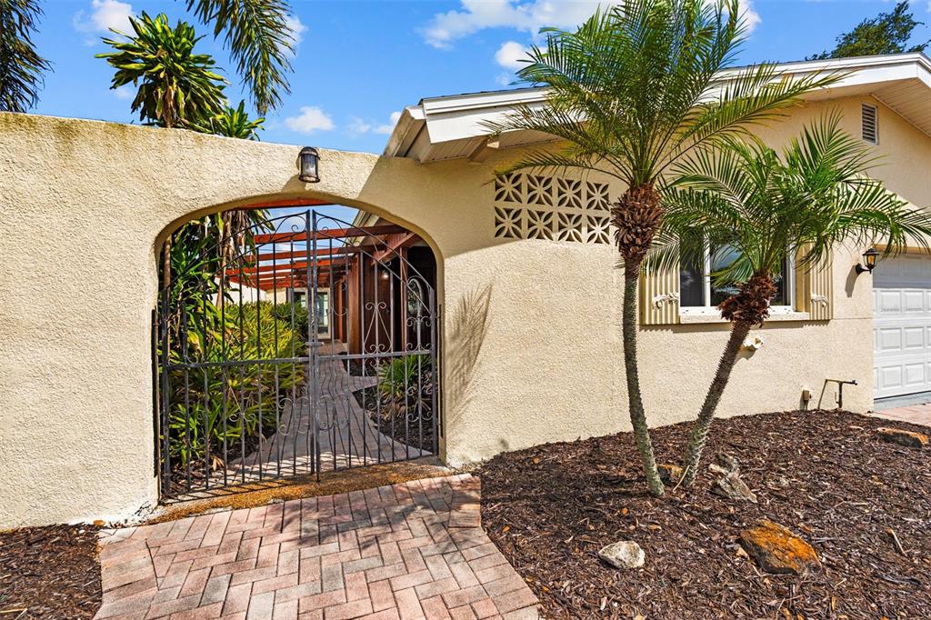 A paved walkway leads to a gated courtyard entrance of a stucco house with palm trees — 10472 ORANGE BLOSSOM LANE, SEMINOLE, FL
