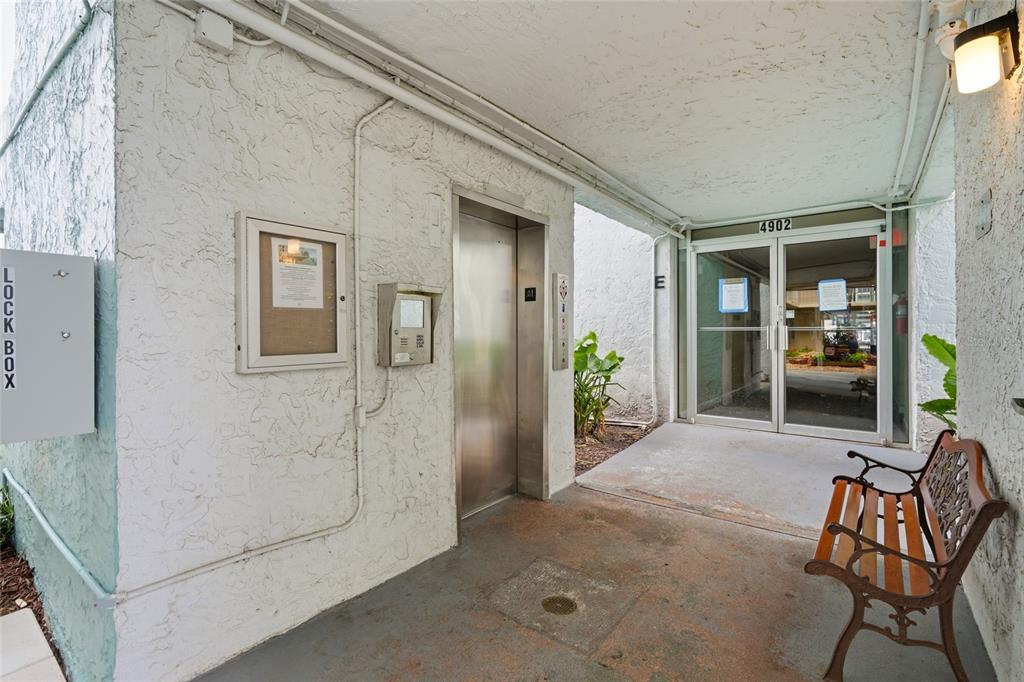 An outdoor building entrance features a metal elevator, glass doors, and a wooden bench — 4900 38TH WAY S UNIT 404, ST PETERSBURG, FL