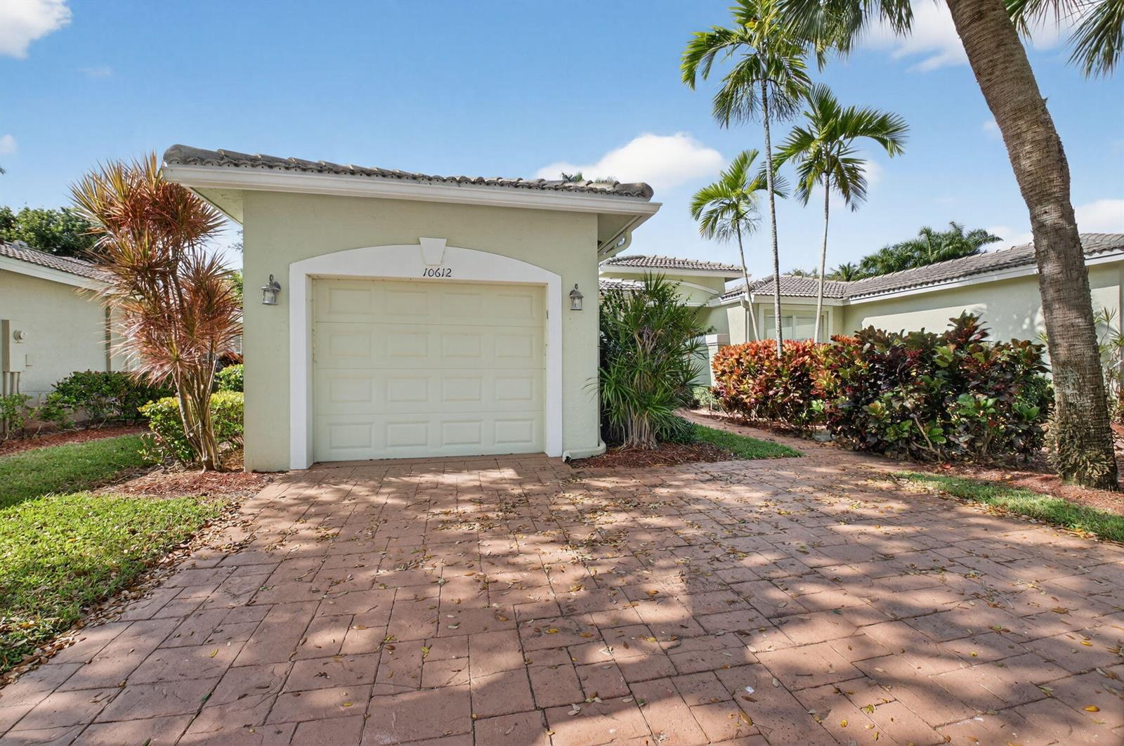 A single-car garage with a paved driveway and tropical landscaping under a blue sky — 10612 Royal Caribbean Circle, Boynton Beach FL 33437, BOYNTON BEACH, FL