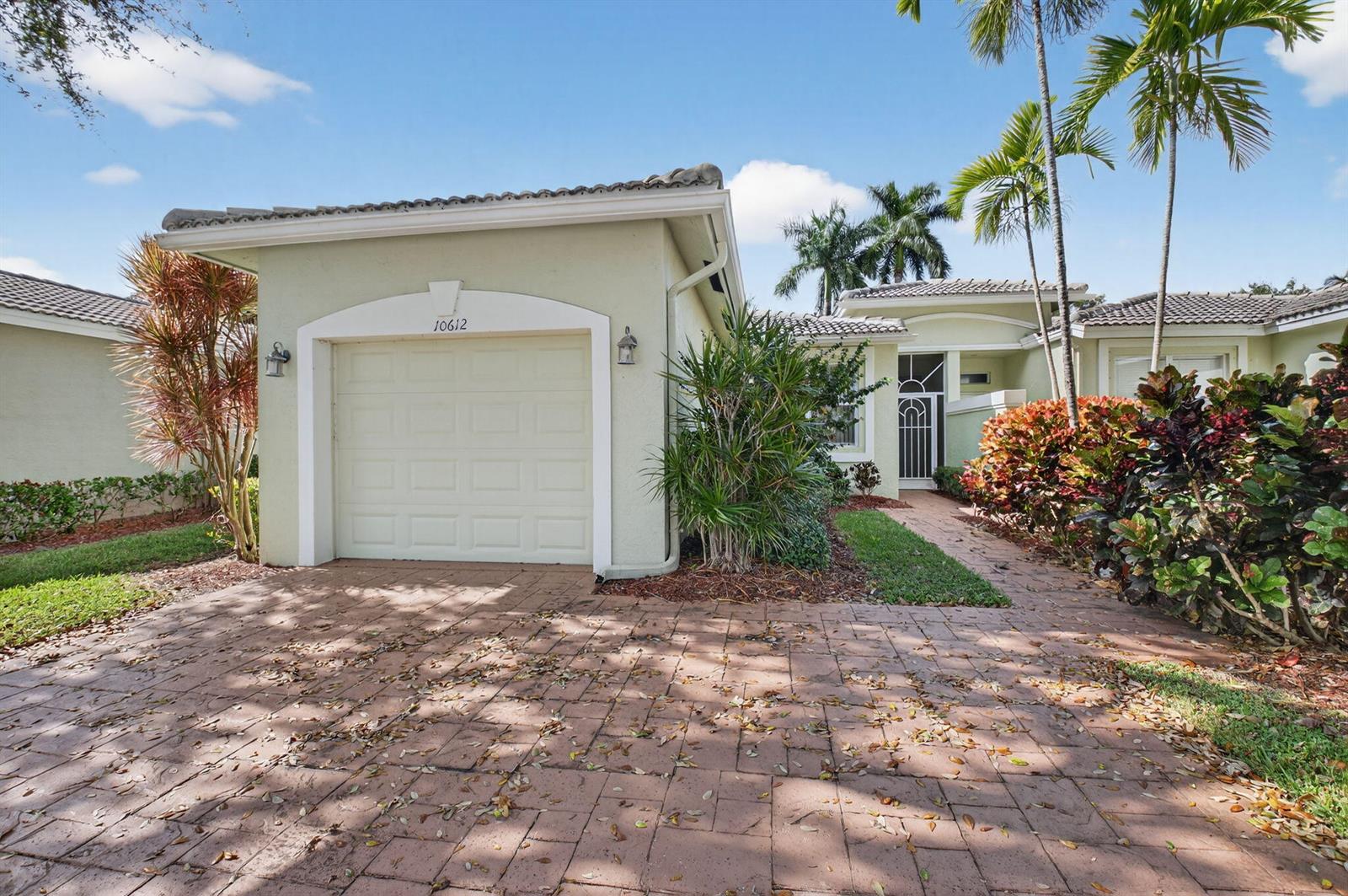 A light green house features a single-car garage, a paved driveway, and tropical landscaping — 10612 Royal Caribbean Circle, Boynton Beach FL 33437, BOYNTON BEACH, FL