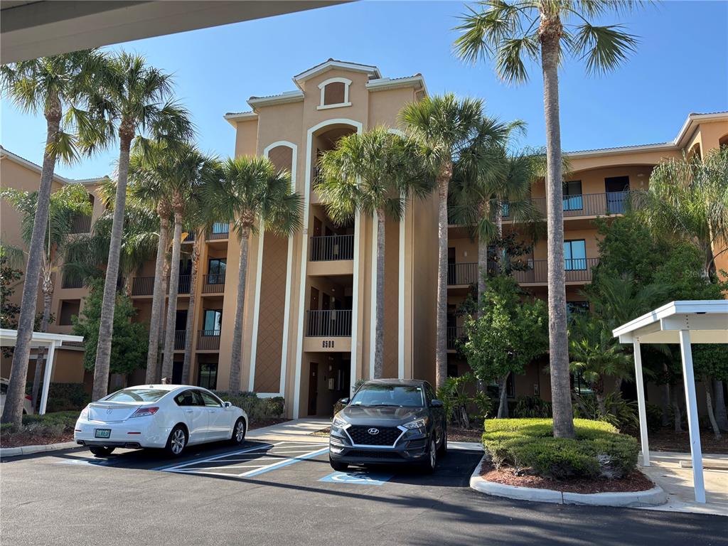 A multi-story tan apartment building with palm trees and parked cars in the foreground — 6509 GRAND ESTUARY TRAIL UNIT 208, BRADENTON, FL