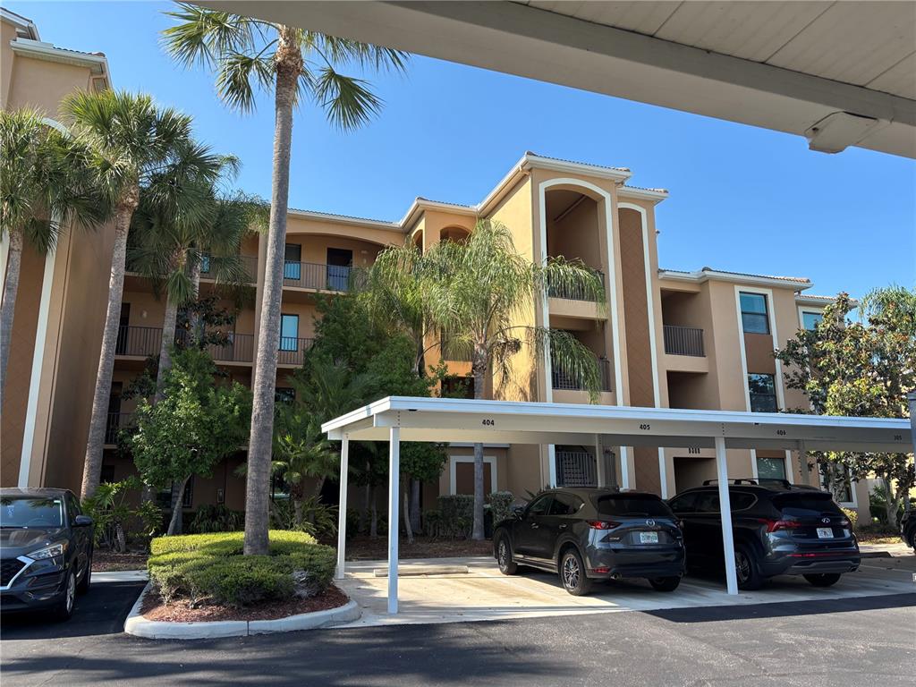 A multi-story tan apartment building features a covered parking area with several parked cars — 6509 GRAND ESTUARY TRAIL UNIT 208, BRADENTON, FL