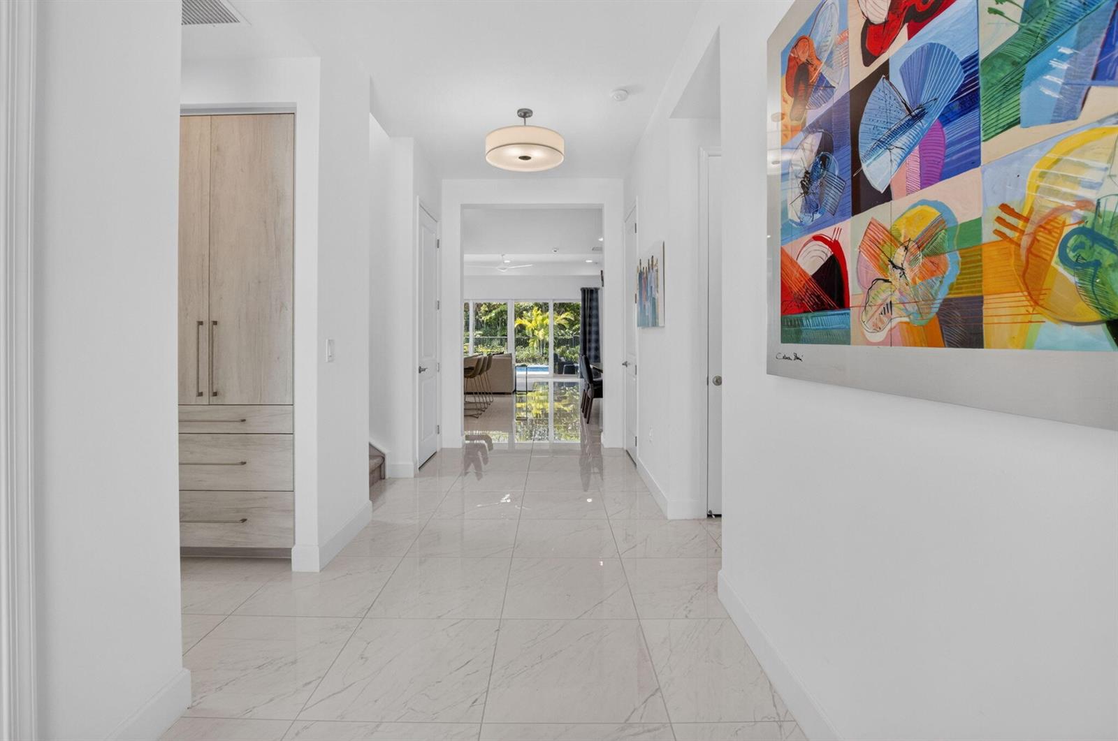 A bright hallway with white walls, light tile floors, a wooden cabinet, and colorful artwork — 13197 Whistler Mountain Road, Delray Beach FL 33446, DELRAY BEACH, FL