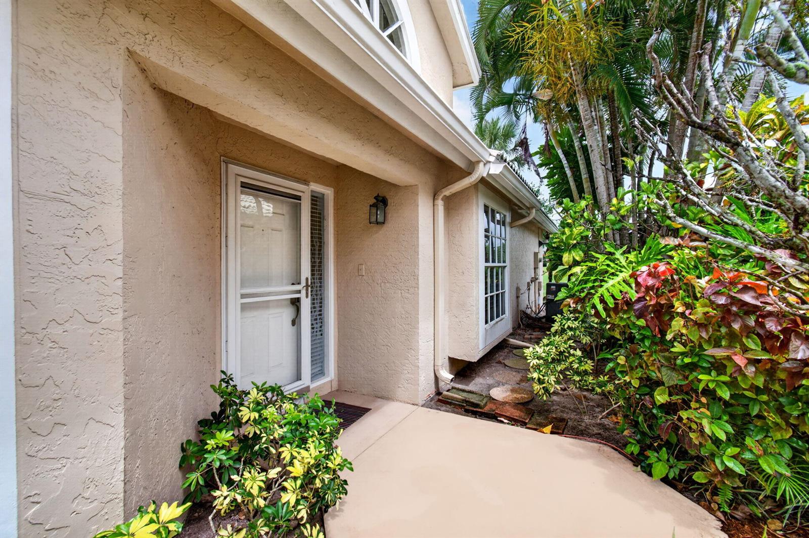 A beige stucco house exterior features a front door with a storm door and landscaping — 6071 Greenspointe Drive, Boynton Beach FL 33437, BOYNTON BEACH, FL