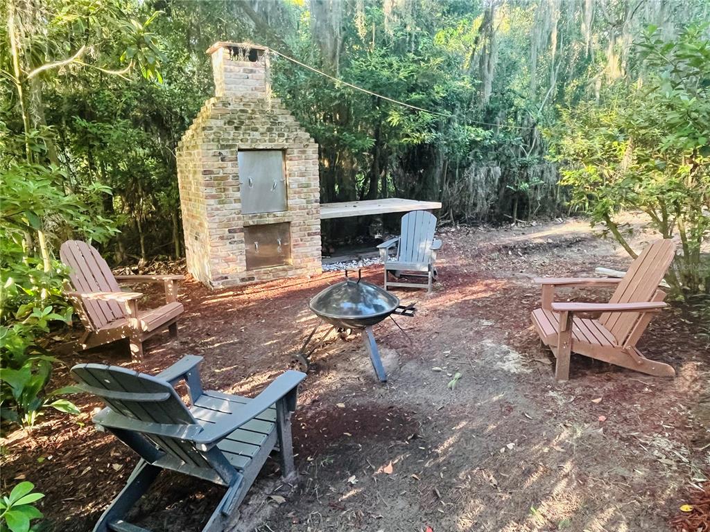 A brick outdoor fireplace and grill area sits in a wooded yard with Adirondack chairs — 1124 NW 4TH STREET NW, GAINESVILLE, FL