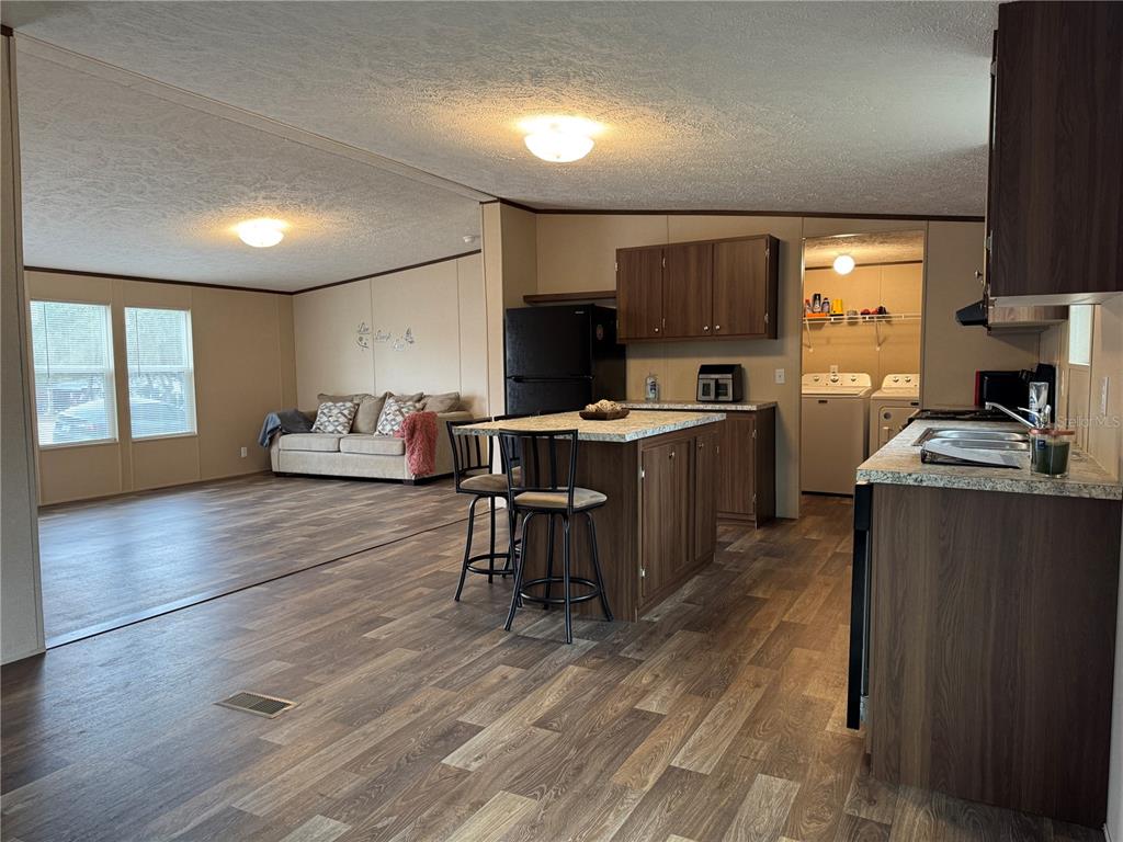 An open-concept kitchen and living area features wood-look flooring, a center island, and dark cabinetry — 18472 SE 58TH PLACE, OCKLAWAHA, FL