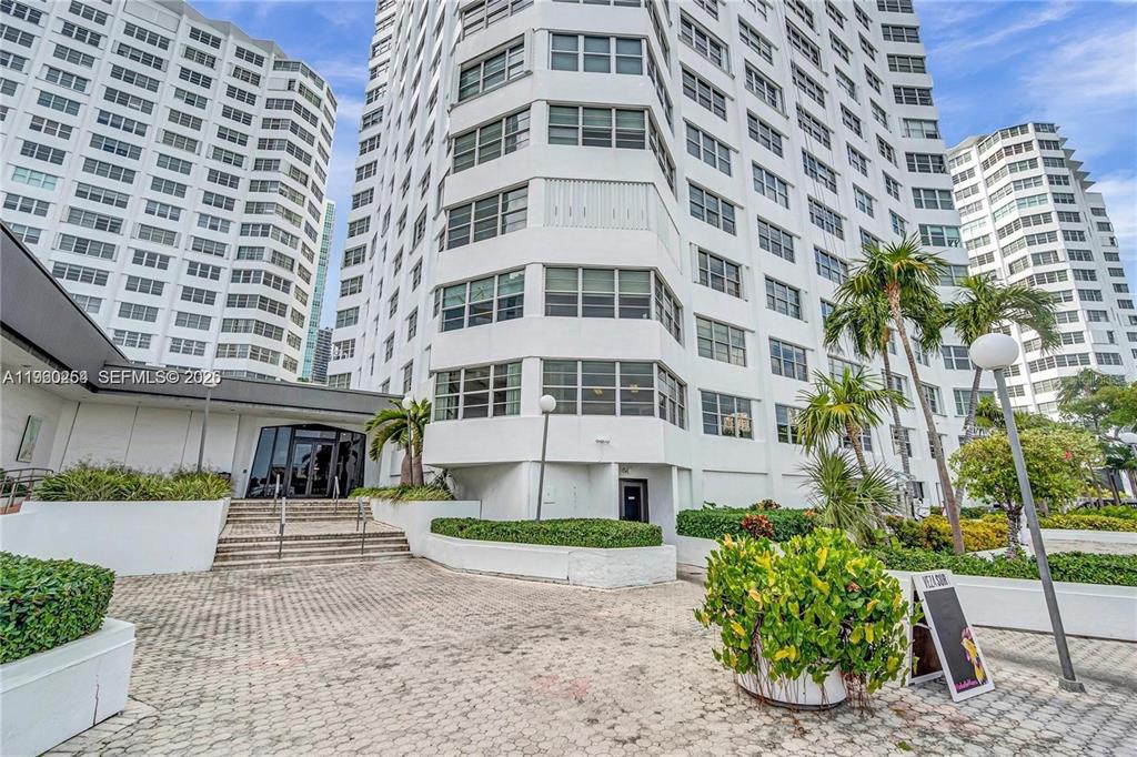 A low-angle exterior view shows the white facade of a multi-story residential apartment building — 999 BRICKELL BAY DRIVE UNIT 1202, MIAMI, FL