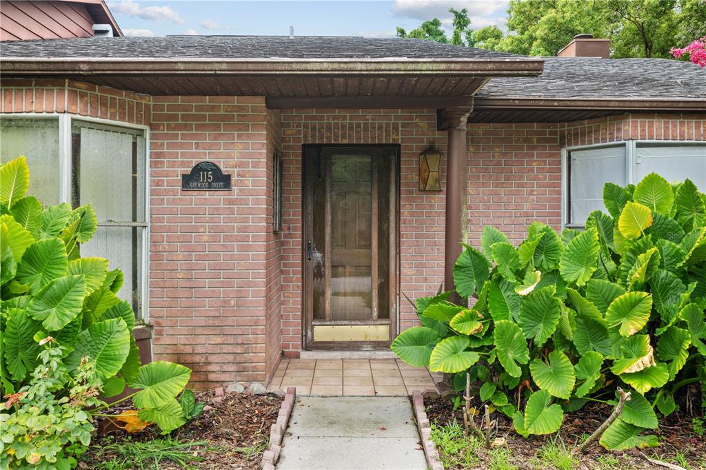 A brick house exterior features a wooden front door, a tiled entryway, and large plants — 115 KAYWOOD DRIVE, SANFORD, FL