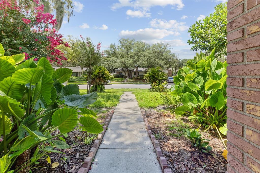 A concrete walkway leads from a brick wall through a garden toward a residential street — 115 KAYWOOD DRIVE, SANFORD, FL