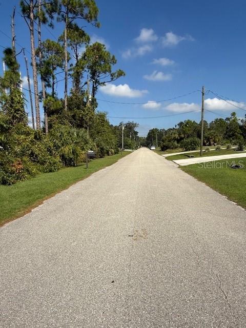 A paved road stretches into the distance, flanked by trees and grassy areas under blue skies — PINCUSHION STREET, NORTH PORT, FL