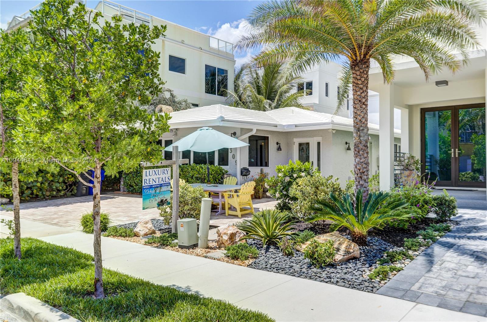 A light-colored house features a patio with yellow chairs, an umbrella, and tropical landscaping — 309 Oak St, Hollywood FL 33019, HOLLYWOOD, FL