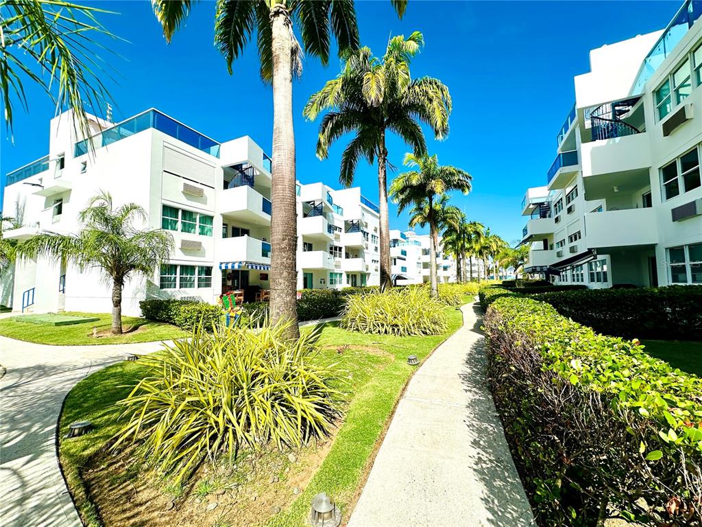 A paved walkway winds through a landscaped courtyard between two white multi-story apartment buildings — COND COSTAMAR BEACH VILLAGE UNIT CO-203, LOIZA, PR