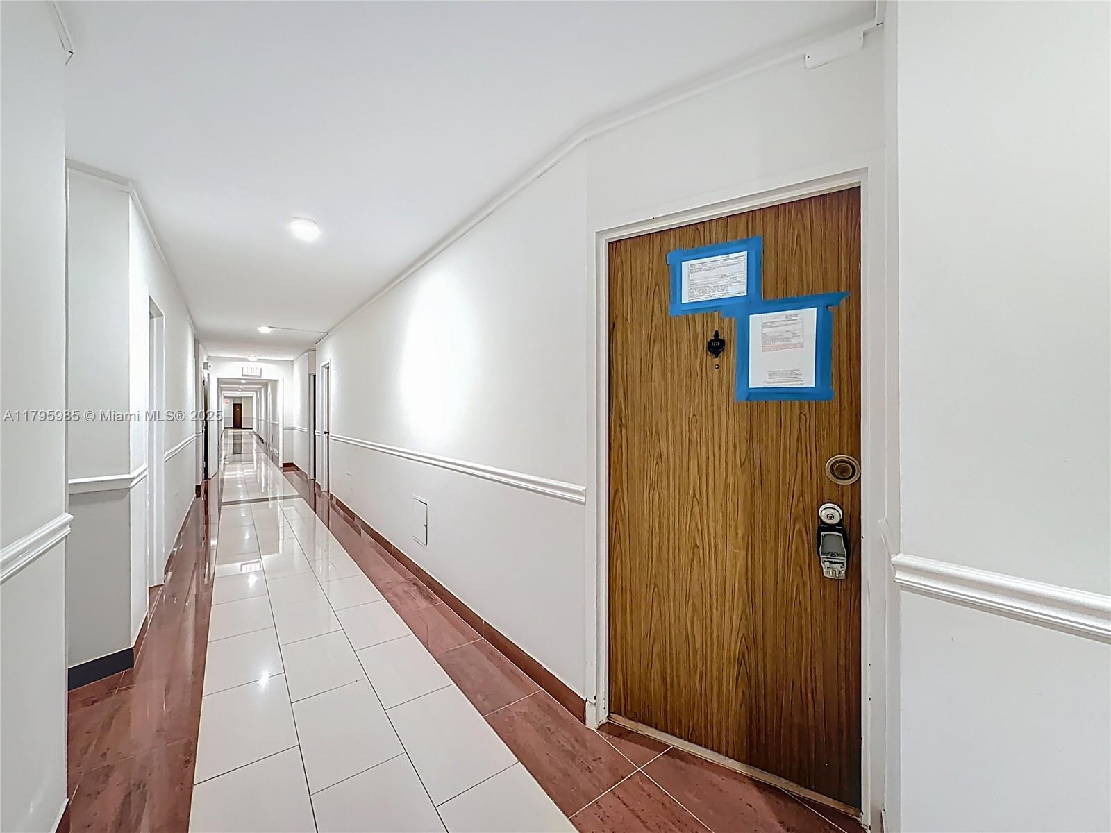 A wooden apartment door with taped notices sits in a long, tiled hallway — 2899 Collins Ave # 1218, Miami Beach FL 33140, MIAMI BEACH, FL