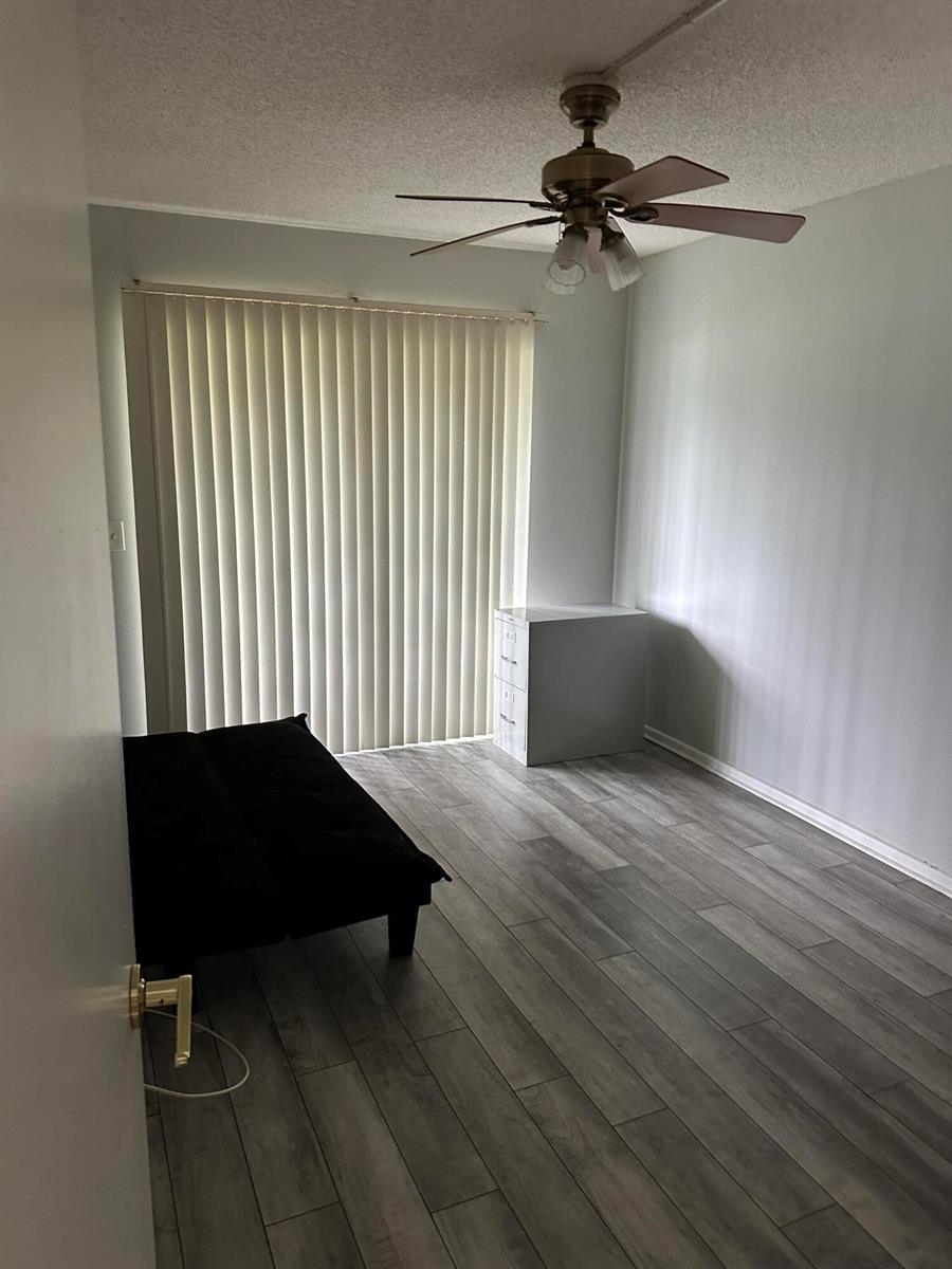 A bedroom with gray wood-look flooring, a black futon, a ceiling fan, and vertical blinds — 599 Fanshaw O, Boca Raton FL 33434, BOCA RATON, FL