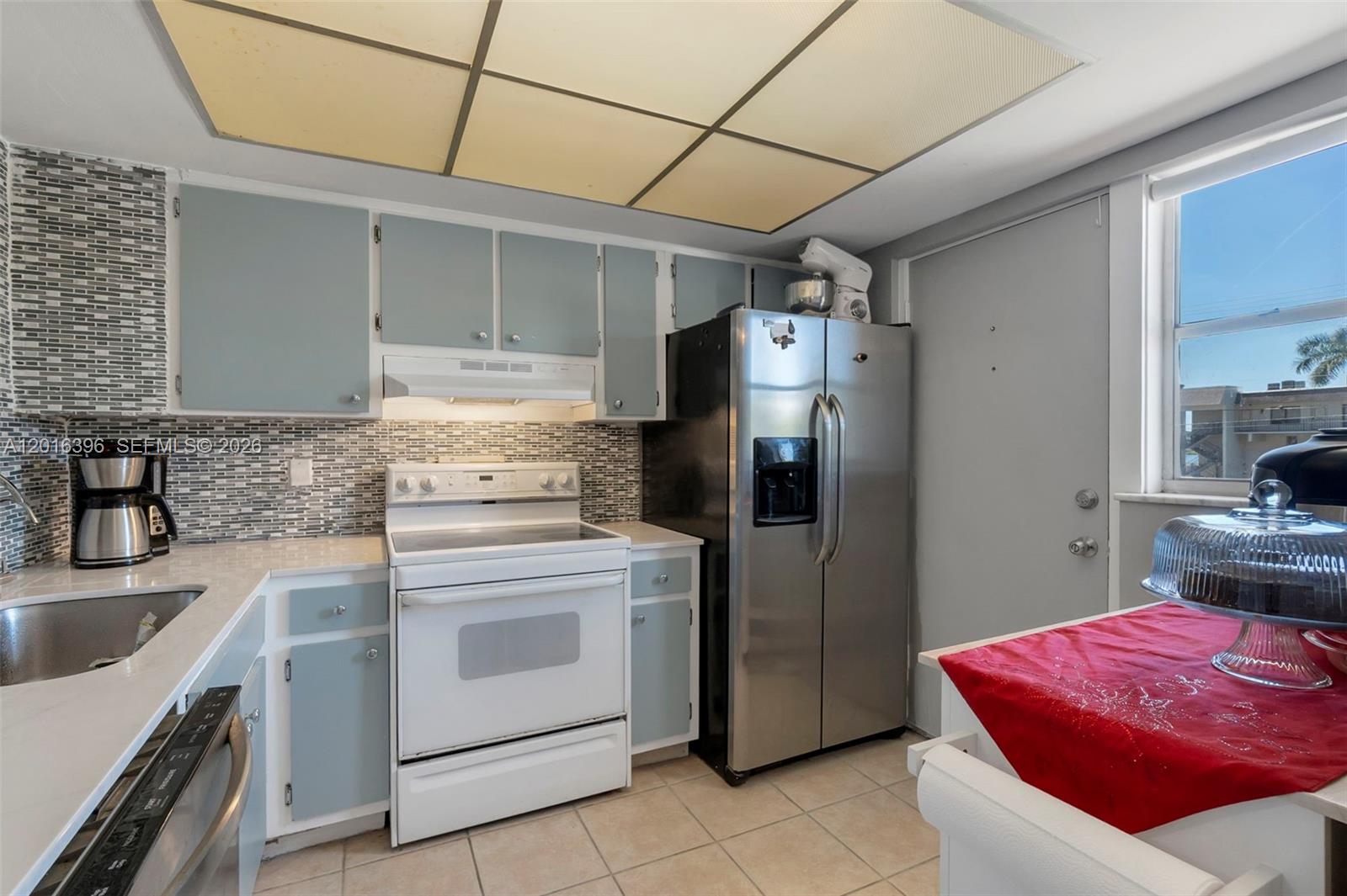 A kitchen features light blue cabinets, a white stove, stainless steel refrigerator, and tiled floors — 1101 Crystal Lake Dr # 203, Deerfield Beach FL 33064, DEERFIELD BEACH, FL