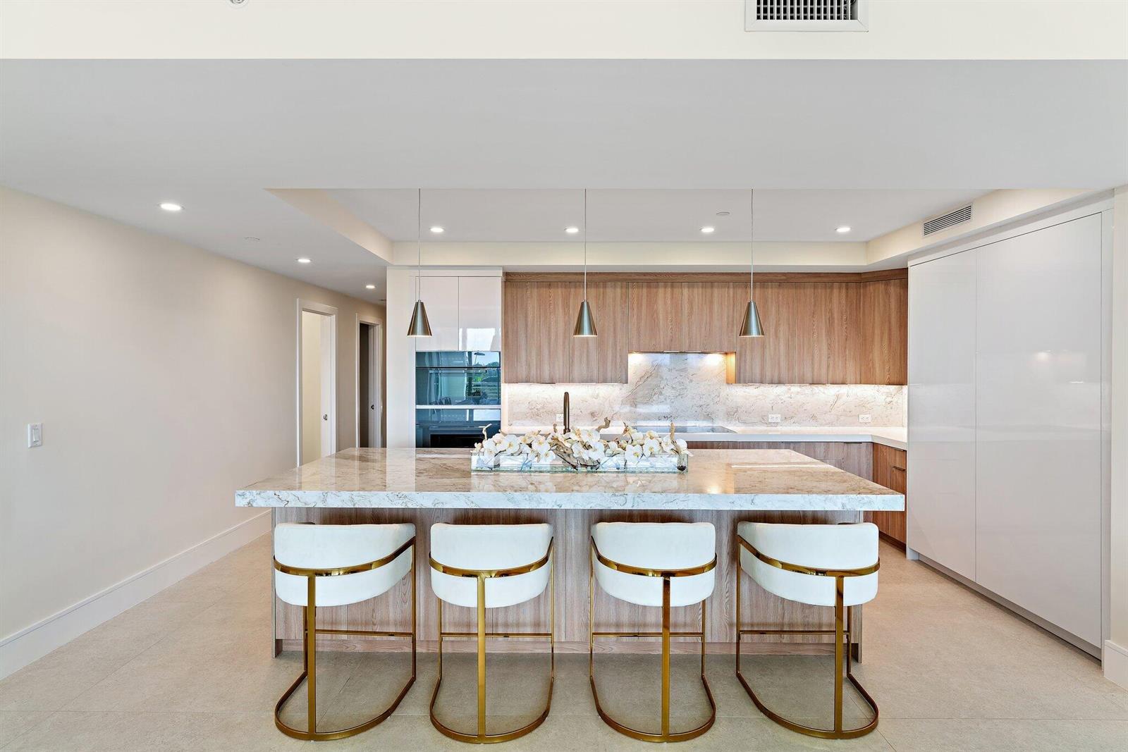 A modern kitchen features a marble island with four gold-accented stools and wood cabinetry — 1 S Coastal Way # 3030, Jupiter FL 33477, JUPITER, FL