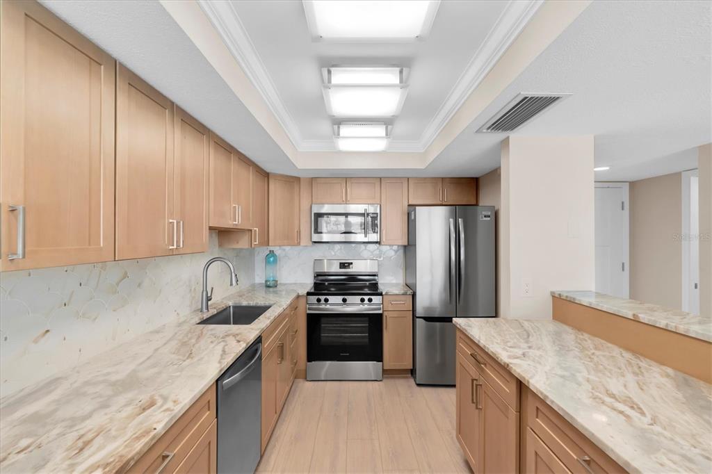 A kitchen features light wood cabinets, stone countertops, stainless steel appliances, and a tiled backsplash — 450 TREASURE ISLAND CAUSEWAY UNIT 102, TREASURE ISLAND, FL