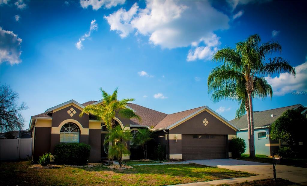 A brown single-story house with a two-car garage and palm trees under a blue sky — 7927 ROUNDELAY DRIVE, NEW PORT RICHEY, FL