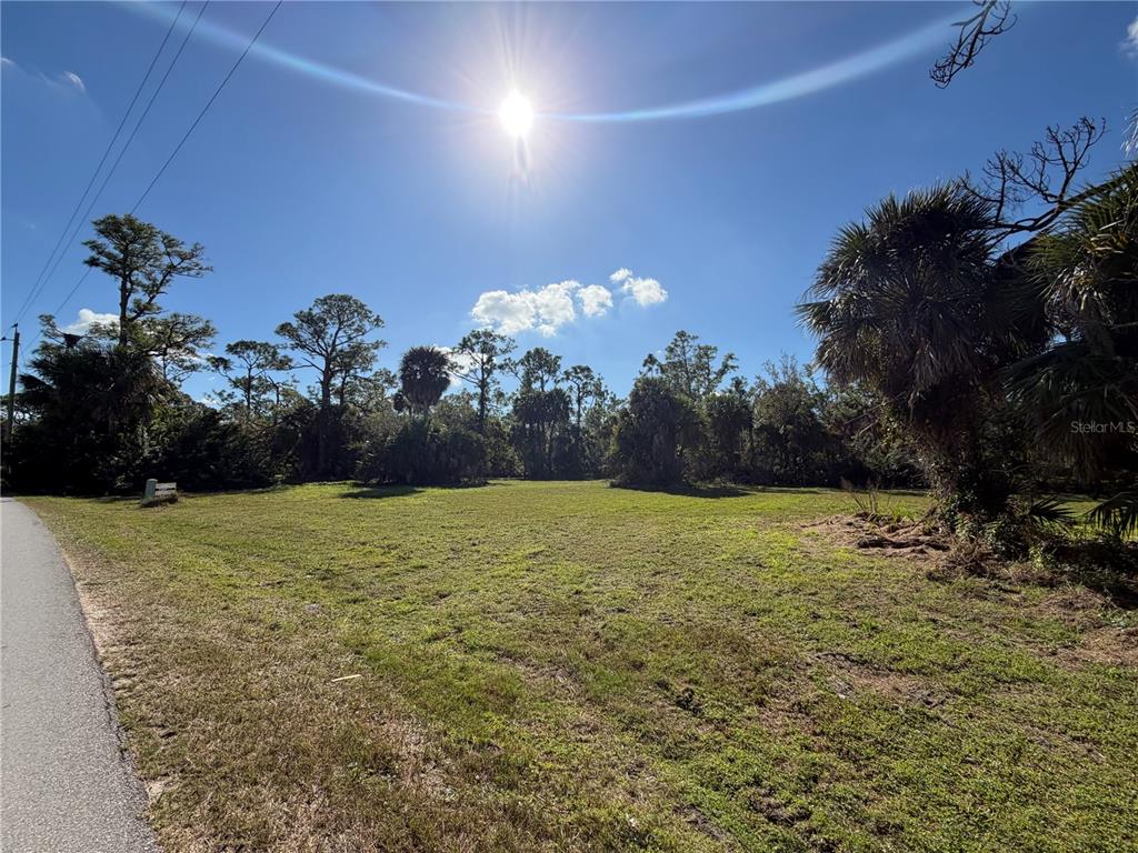 A grassy, vacant lot is bordered by trees and a paved road under a sunny sky — 24501 YACHT CLUB BOULEVARD, PUNTA GORDA, FL