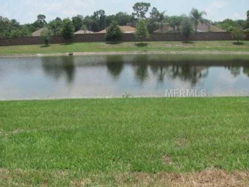 A grassy lawn overlooks a calm pond with a brick wall and trees in the background — 3809 BLACK SPRUCE LANE, WINTER SPRINGS, FL