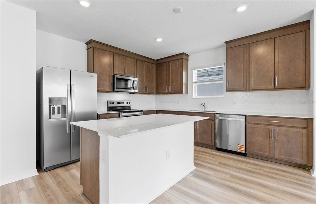 A modern kitchen features brown wood cabinets, stainless steel appliances, and a white island countertop — 1452 HAMILTON WAY, LAKE ALFRED, FL