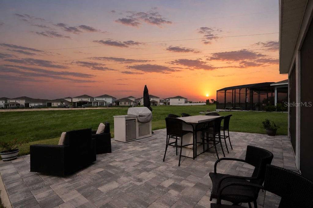 A paved patio features outdoor furniture and a grill overlooking a grassy field at sunset — 1193 WHISPERING LANE, MINNEOLA, FL