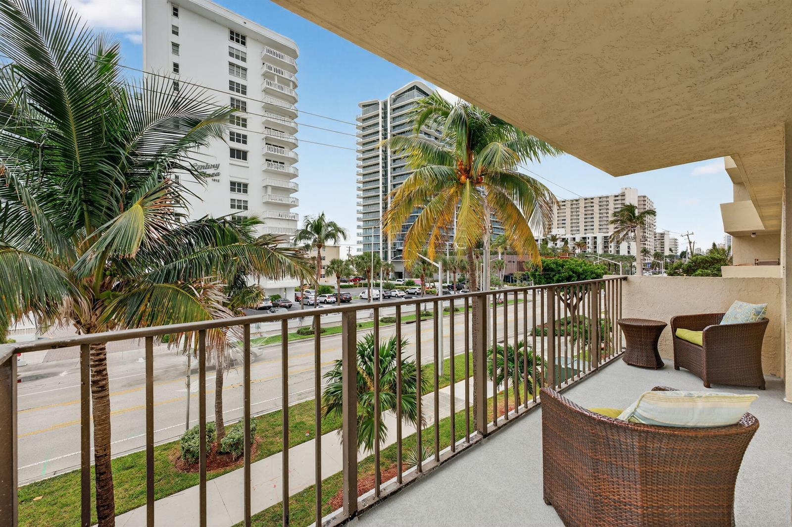 A balcony with wicker chairs overlooks a street lined with palm trees and high-rise buildings — 1009 N Ocean Boulevard # 312, Pompano Beach FL 33062, POMPANO BEACH, FL