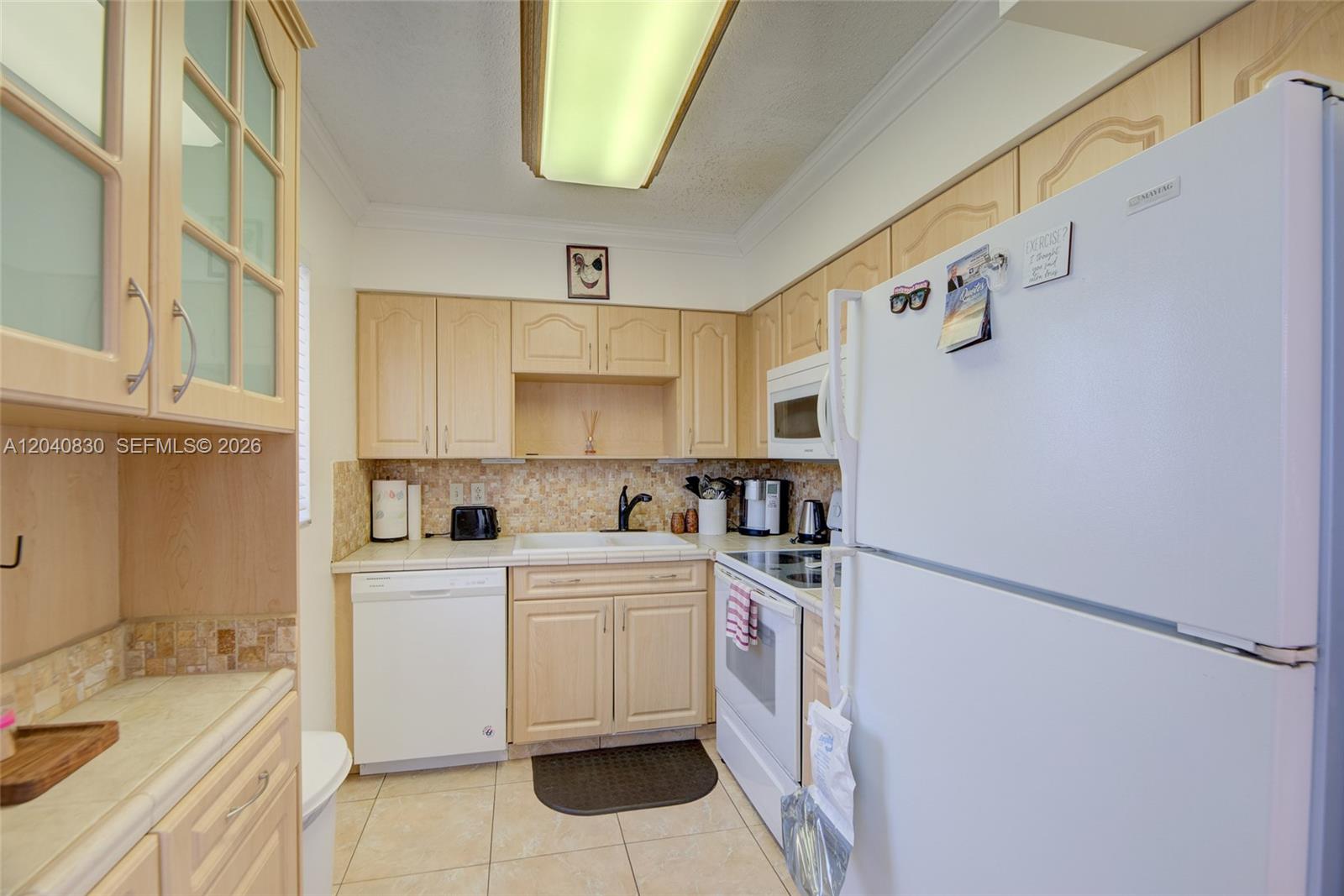 A kitchen features light wood cabinets, white appliances, tiled countertops, and a beige tile floor — 920 SW 10th Ter # 12R, Hallandale Beach FL 33009, HALLANDALE BEACH, FL