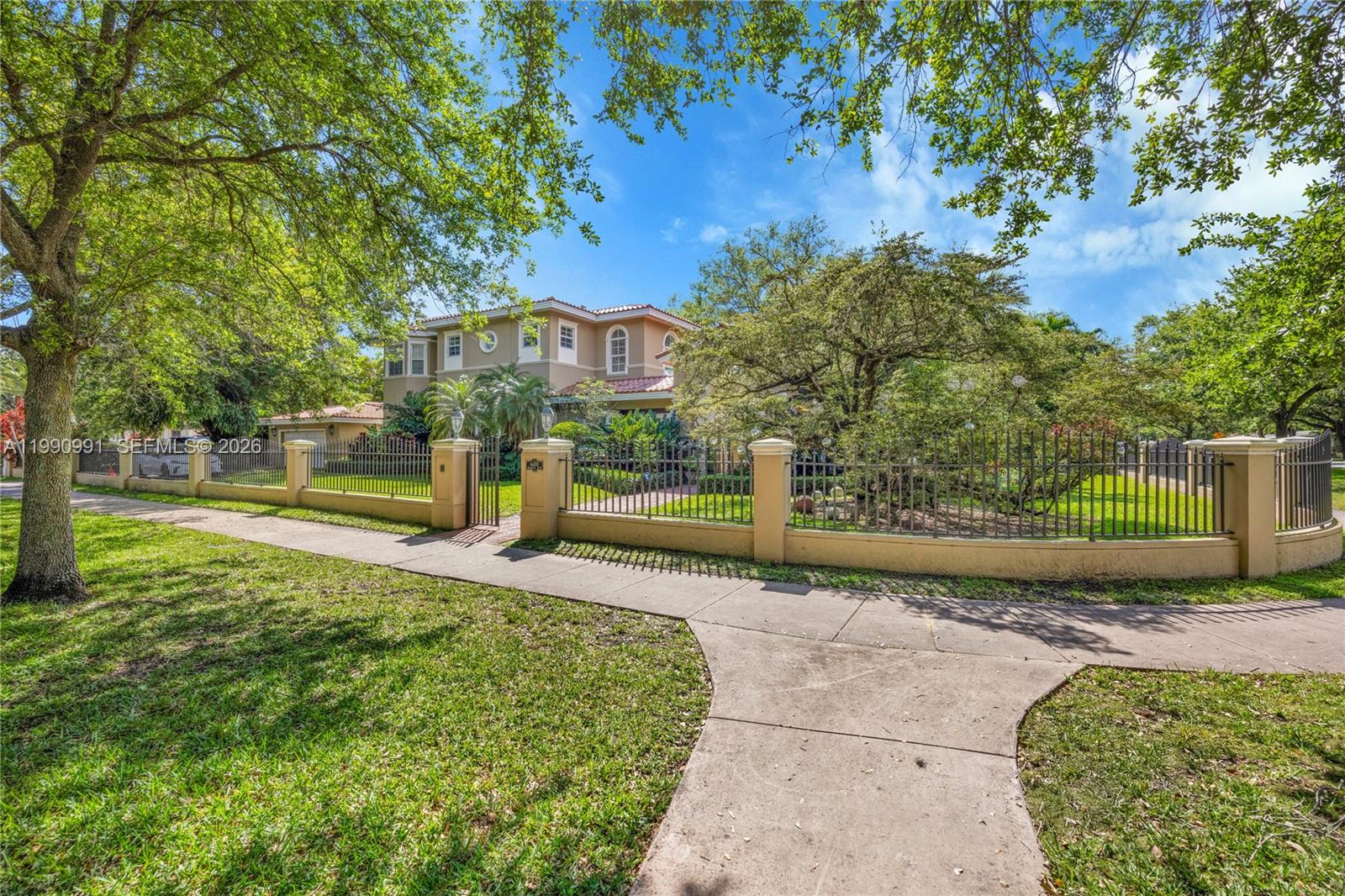 A two-story house with a fenced yard and mature trees is viewed from the sidewalk — 6200 Granada Blvd, Coral Gables FL 33146, CORAL GABLES, FL