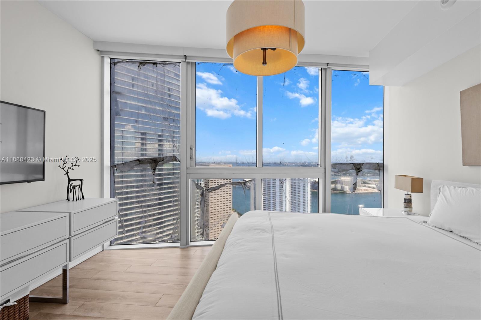 A modern bedroom features a white bed, a dresser, and floor-to-ceiling windows overlooking a city — 465 Brickell Ave # 4901, Miami FL 33131, MIAMI, FL
