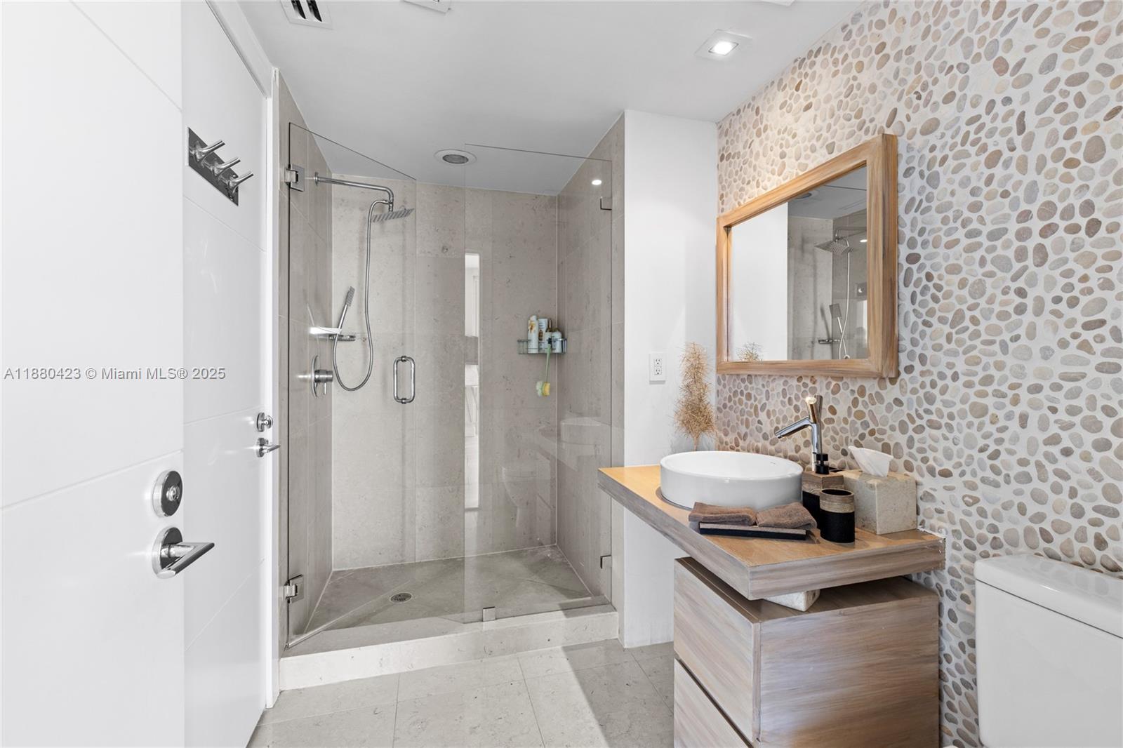 A modern bathroom features a glass-enclosed shower, a pebble-tiled wall, and a wooden vanity — 465 Brickell Ave # 4901, Miami FL 33131, MIAMI, FL