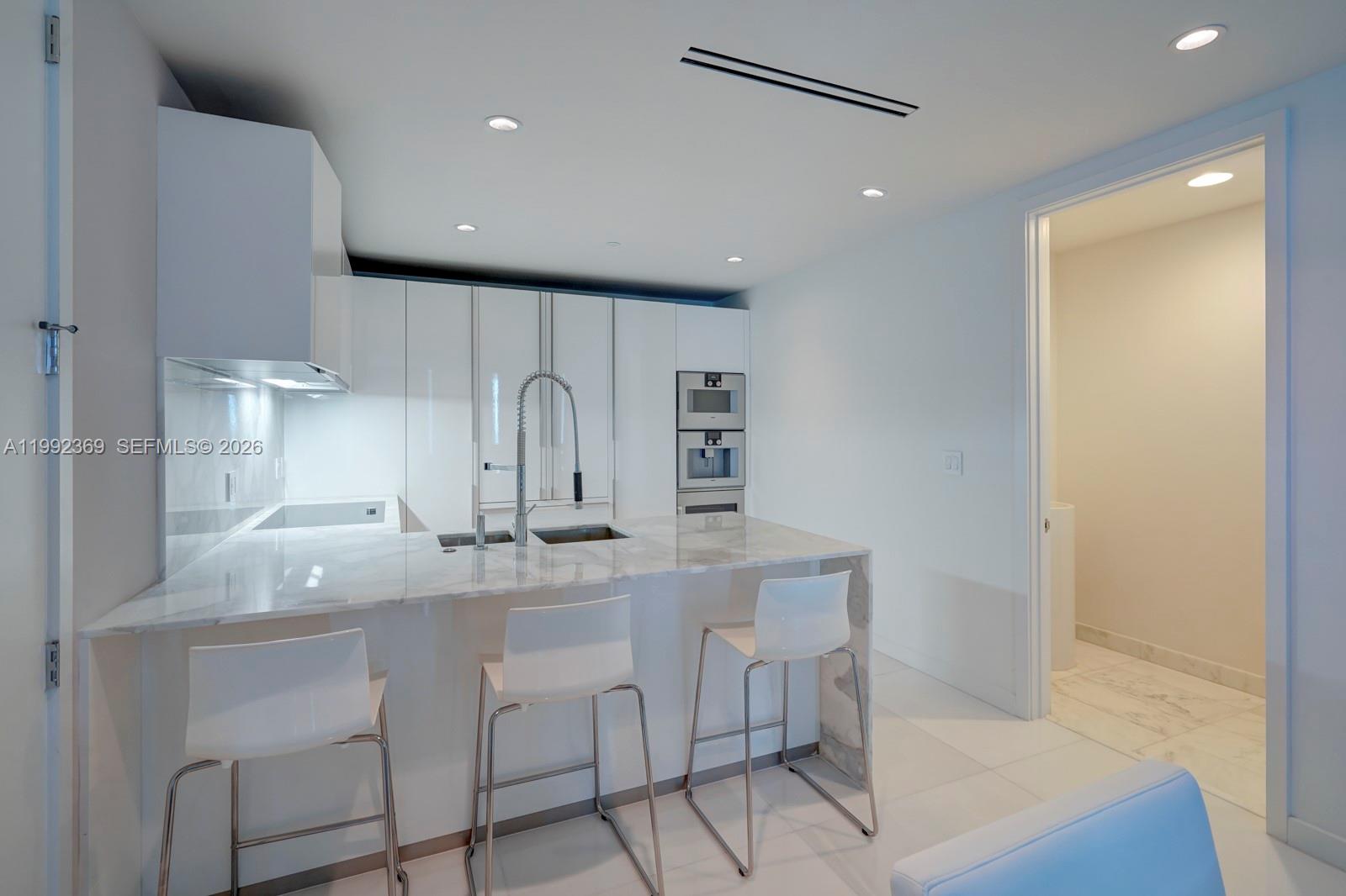 A modern kitchen features white cabinetry, a marble-topped island with bar stools, and stainless appliances — 10203 Collins Ave # 1005, Bal Harbour FL 33154, BAL HARBOUR, FL