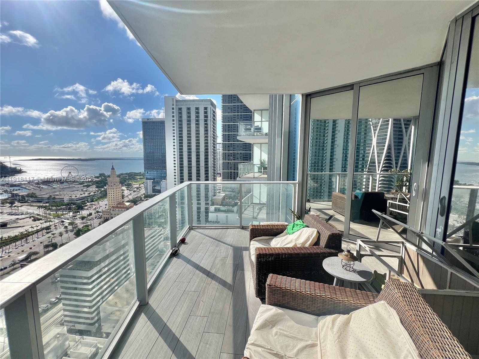 A high-rise balcony features wicker chairs, a small table, and views of the city skyline — 851 NE 1st Ave # 2605, Miami FL 33132, MIAMI, FL