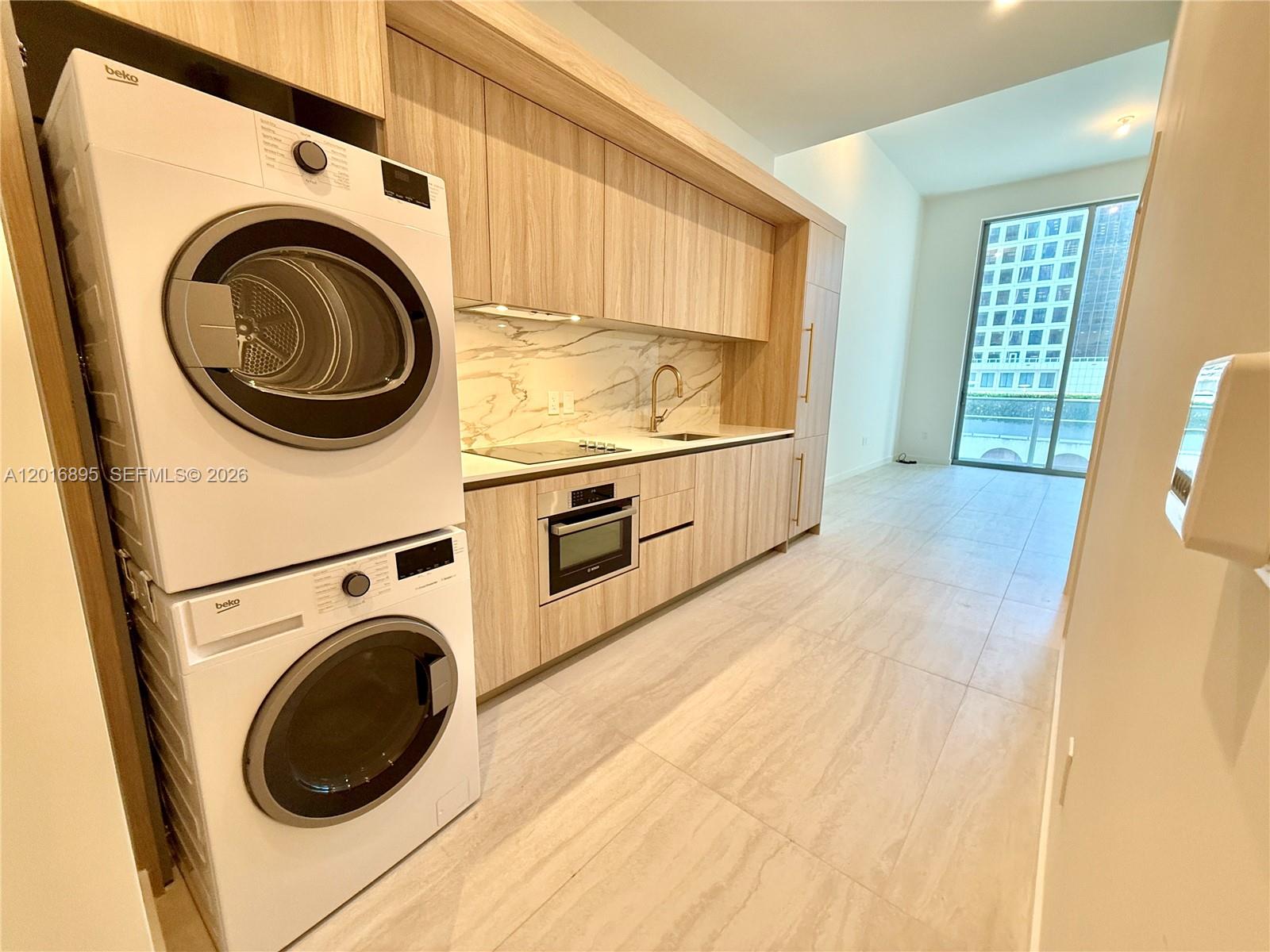 A modern kitchen features light wood cabinetry, marble backsplash, and a stacked washer and dryer — 77 SE 5 st # 1213, Miami FL 33131, MIAMI, FL