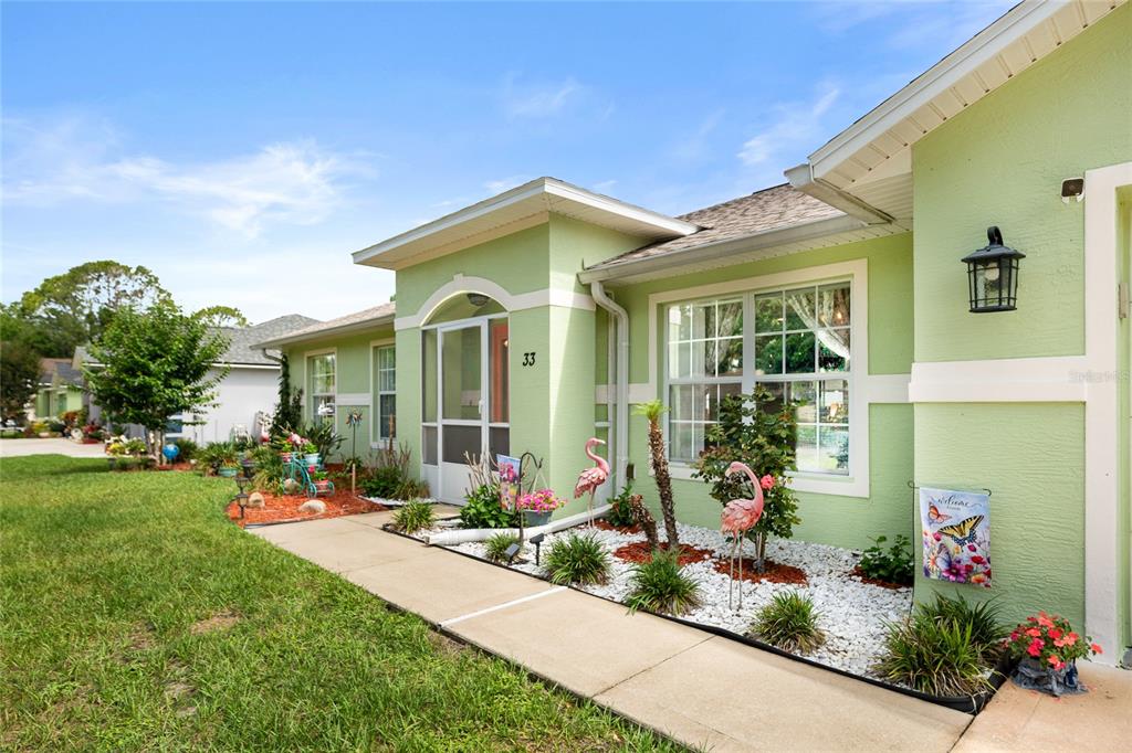 A light green house exterior features a front walkway, white rock landscaping, and flamingo decorations — 33 ROYAL LEAF LANE, PALM COAST, FL