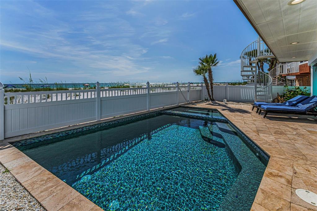 A swimming pool sits on a stone patio next to a white fence and beach — 13303 GULF LANE, MADEIRA BEACH, FL