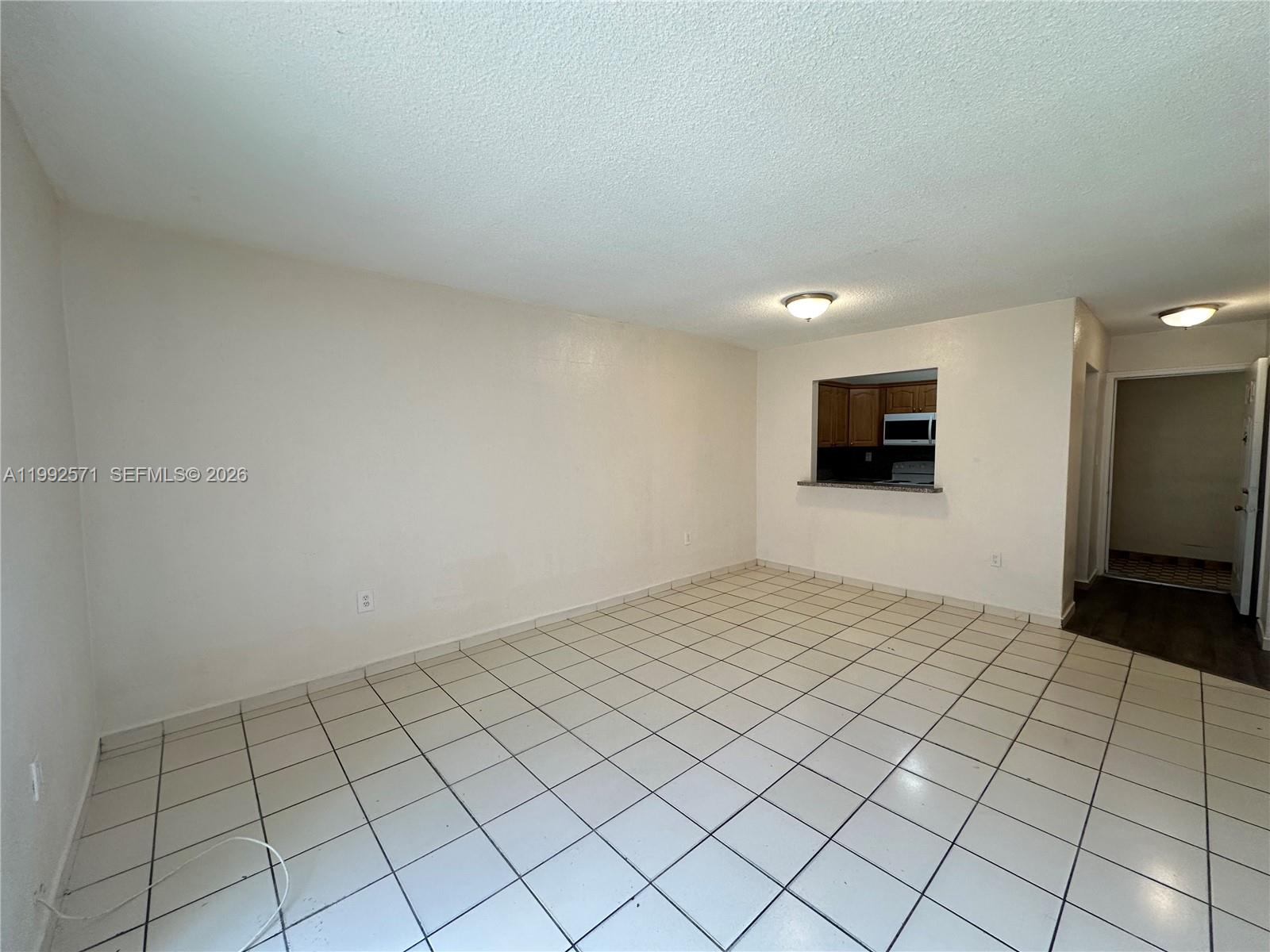 An empty room features white tile flooring, plain walls, and a kitchen pass-through window — 1008 NW 45th Ave # 08, Miami FL 33126, MIAMI, FL