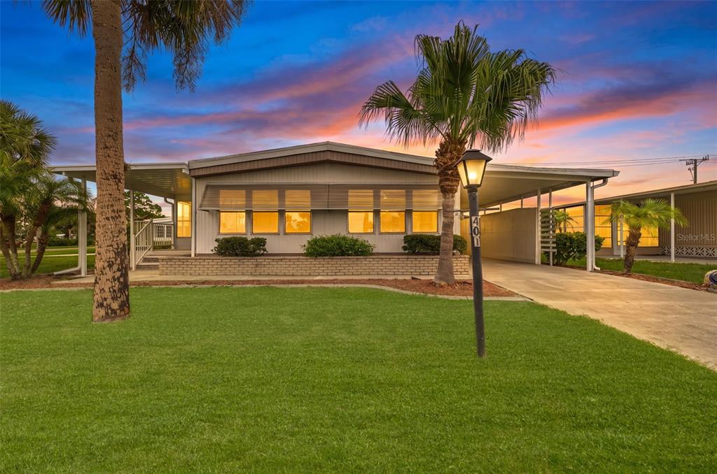 A single-story manufactured home with a carport, palm trees, and a lawn at sunset — 401 LOMA LINDA, NORTH PORT, FL