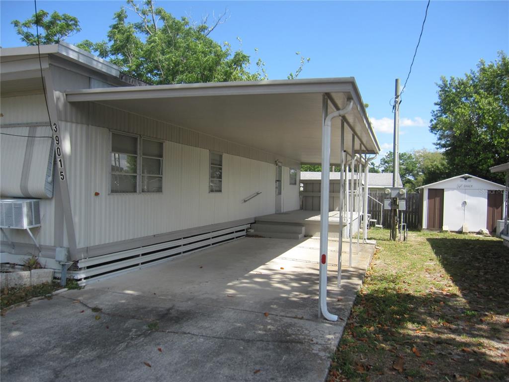 A mobile home exterior features a concrete carport, a covered porch, and a small shed — 39015 HILLCREST DRIVE, ZEPHYRHILLS, FL