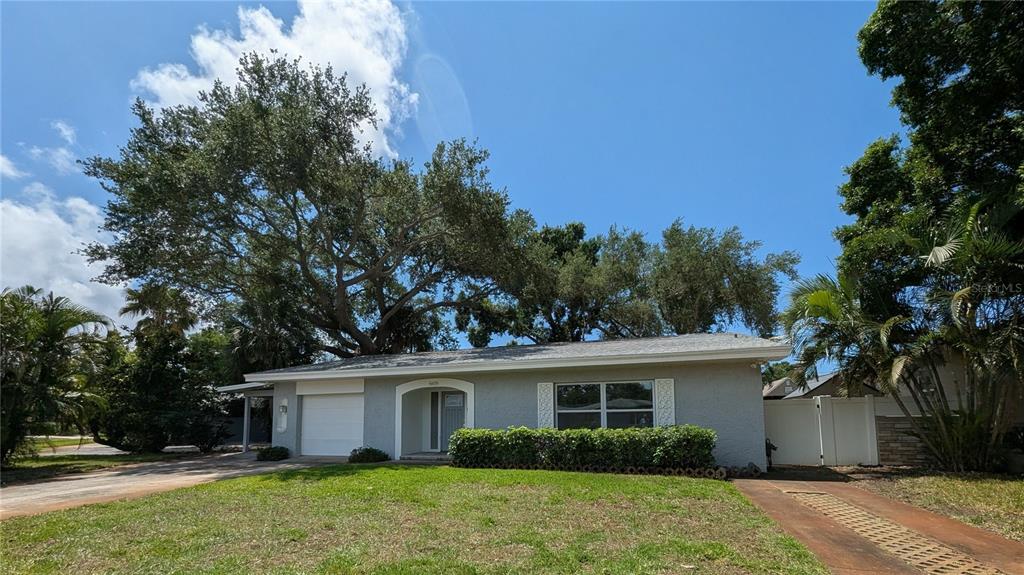 A single-story gray house with a white garage door sits behind a large oak tree — 6408 7TH AVENUE N, ST PETERSBURG, FL
