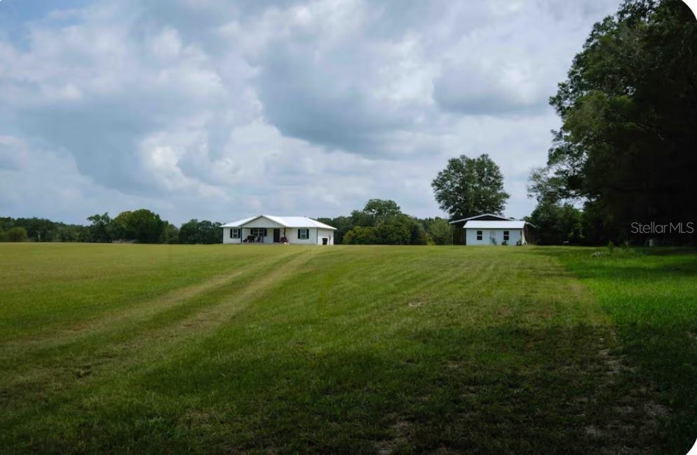A wide, grassy field leads to a white house and a separate outbuilding under clouds — 21411 NW 209TH TERRACE, HIGH SPRINGS, FL