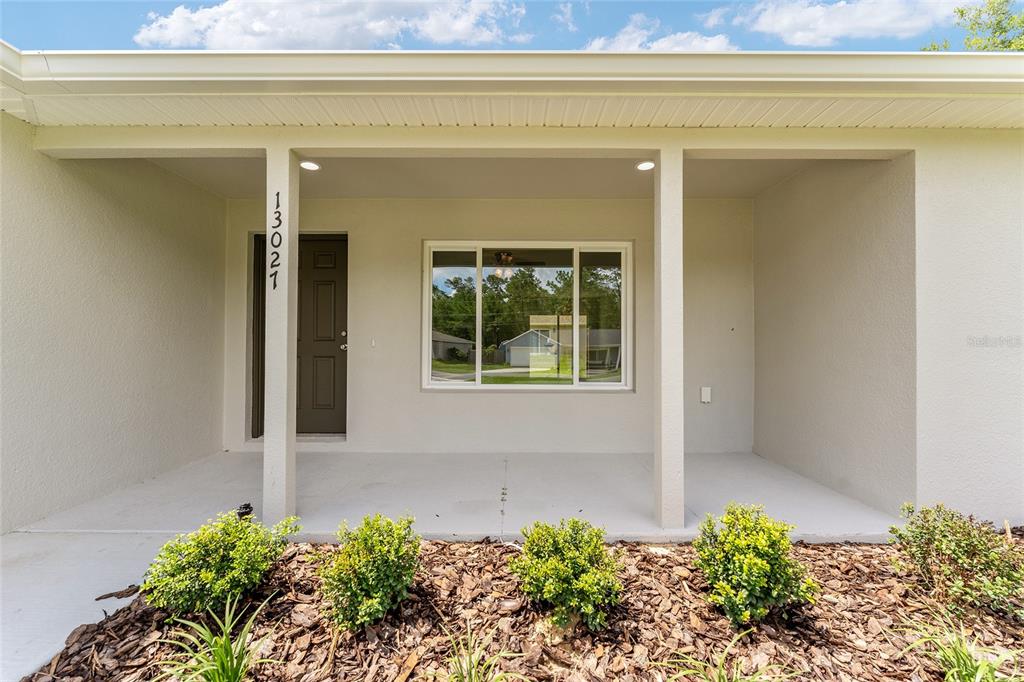 A covered front porch features a dark door, a large window, and landscaped mulch beds — 13027 NE 5TH PLACE, SILVER SPRINGS, FL