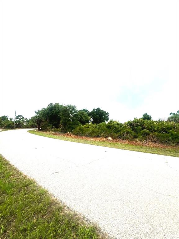 A paved road curves past a grassy area and dense vegetation under a bright sky — 7441 MANVILLE TERRACE, PORT CHARLOTTE, FL