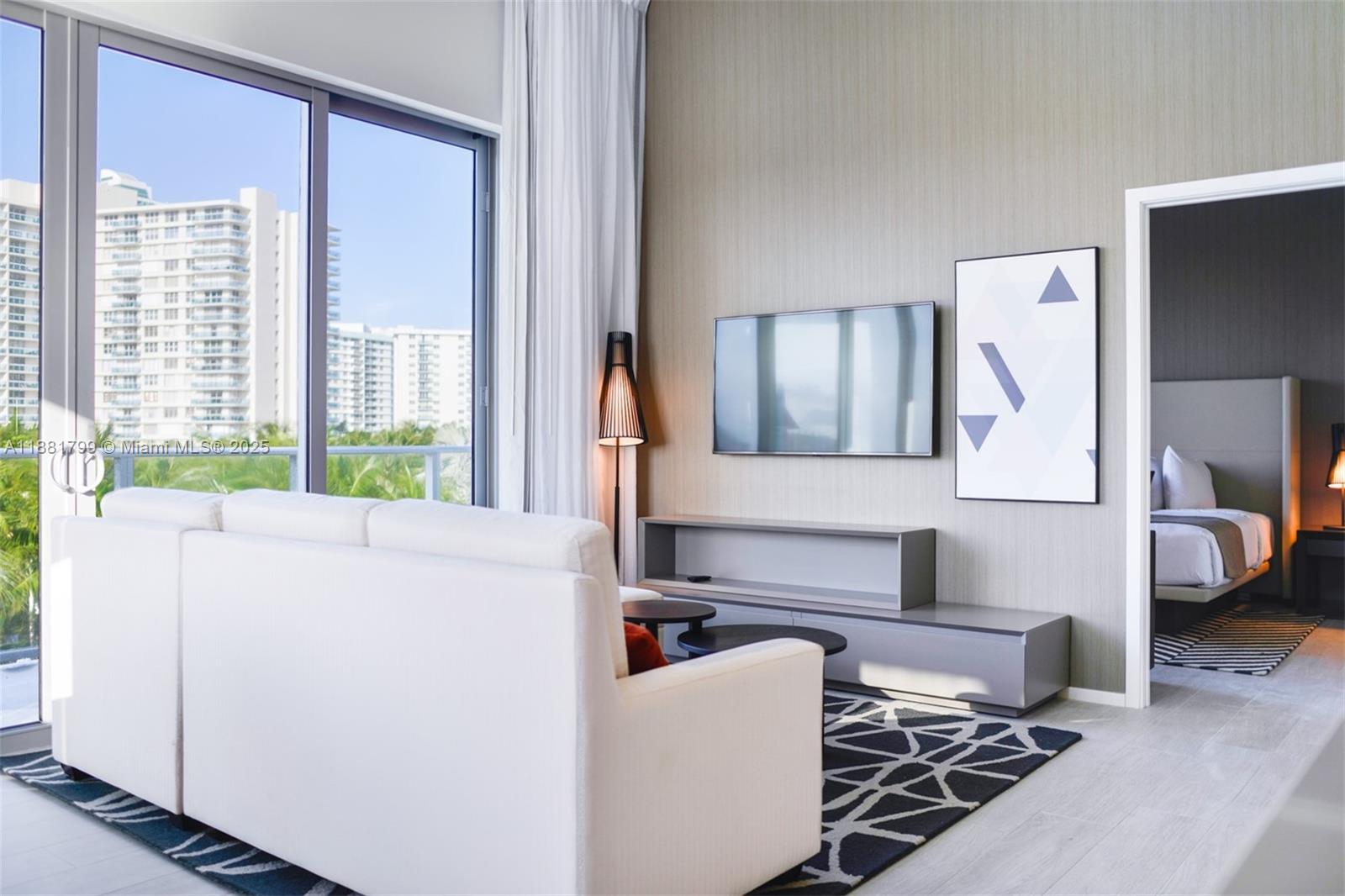 A modern living room features a white sofa, a wall-mounted television, and a bedroom view — 4010 S Ocean Dr # R302, Hollywood FL 33019, HOLLYWOOD, FL