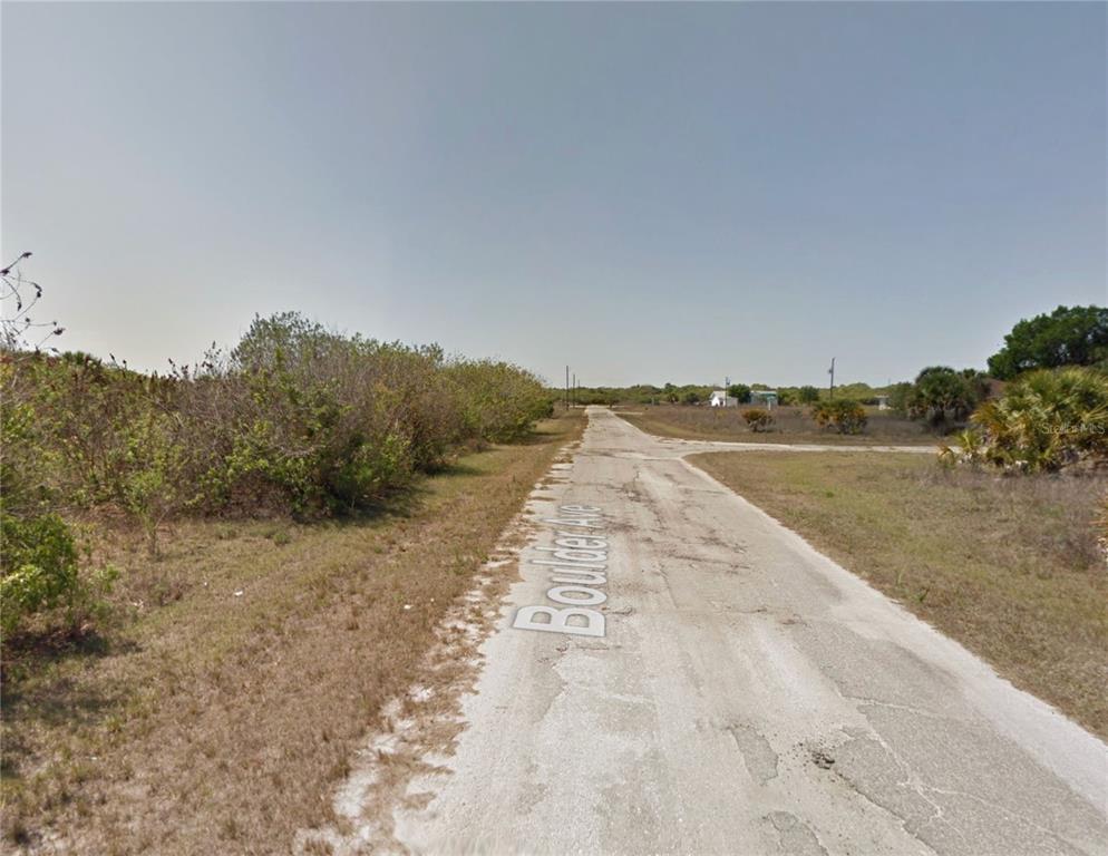 A narrow, weathered paved road stretches through an undeveloped, grassy area with scattered shrubs — 19425 BOULDER AVENUE, PORT CHARLOTTE, FL